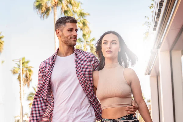 Trendy attractive couple walking together on the street with palms. Beautiful brunette woman with handsome italian man