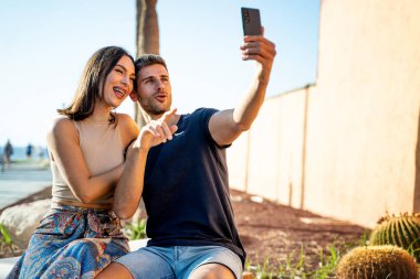 Genç mutlu çift cep telefonuyla selfie çekiyor, akıllı telefona gülümsüyor. Yaz zamanı. Tatilde. Gezgin. Gerçek insan yaşam tarzı..