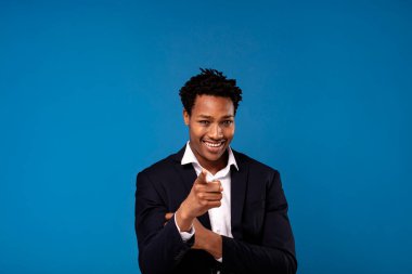 Gülümseyen başarılı Afrikalı iş adamı mavi stüdyo arka planında kamerayı işaret ederek poz veriyor. Bir sürü kopyalama alanı.