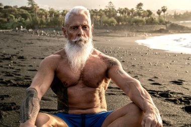 Sağlıklı bir bedende, sağlıklı bir zihinde. Beyaz sakallı ve dövmeli yaşlı adam gün batımında sahilde meditasyon yapıyor. Yaz tatili. Yaşlılar emeklilik kavramında sağlıklı bir yaşam tarzına sahipler