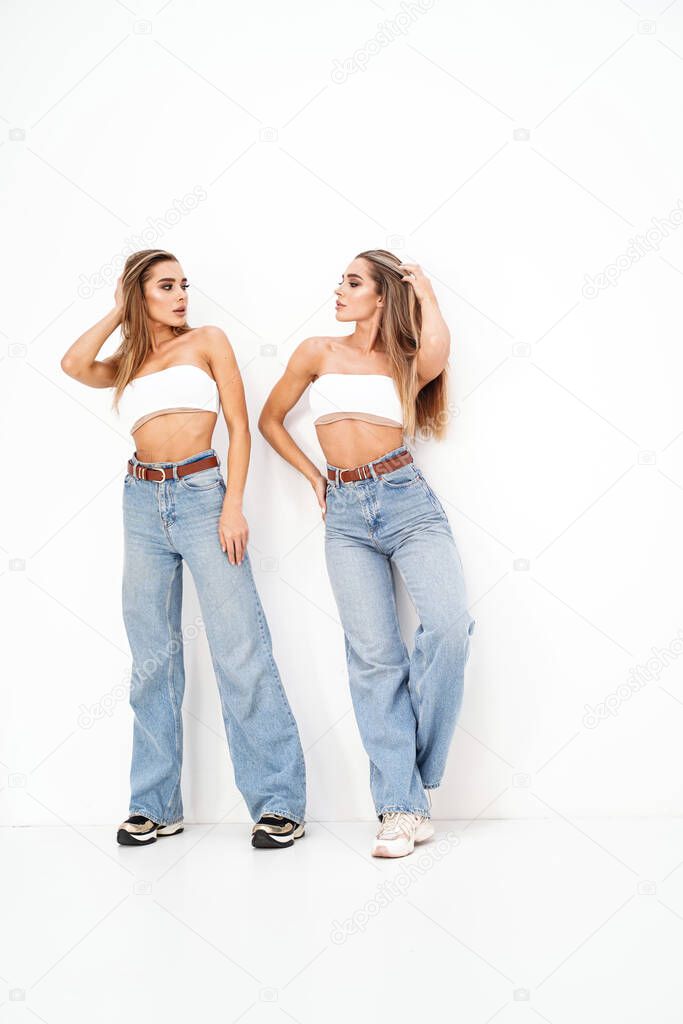 Two beautiful young women with long blond hair and glamour makeup wearing fashionable jeans, standing over white studio background. Sisters. Twins. Studio shot. Copy space
