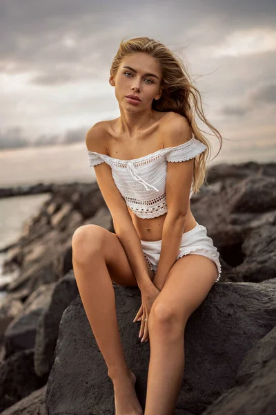 白人金发女子在岩石海滩上休息 性感的女孩看着摄像机坐在岩石上夏天的感觉 — 图库照片