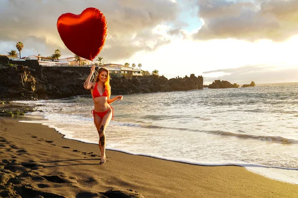 在美丽的海滩上 穿着红色比基尼的快乐的年轻女人拿着大大的心气球微笑 日落时间 情人节 浪漫的约会爱 真实的人的情感 — 图库照片