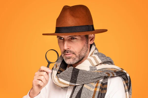 Male detective with magnifying glass, wearing fashionable scarf and brown hat, posing on yellow studio background. Portrait of handsome man with beard.