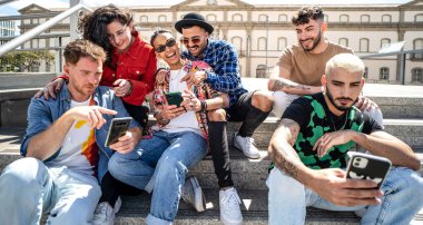 Mutlu bir grup çok ırklı öğrenci, açık havada akıllı telefon kullanan arkadaşlar, sosyal medyayı izlemek ve gülmek, birlikte eğlenmek..