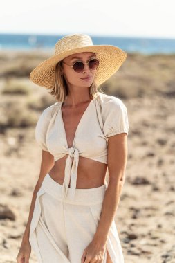 Hasır şapkalı, güneş gözlüklü, şık yaz kıyafetleri giyen, vahşi sahilde poz veren mutlu boho kadının portresi. Güneşli bir gün. Rahatla..