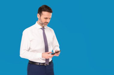 Akıllı telefondan mesaj yazan yakışıklı bir işadamı mavi stüdyo arka planında izole edilmiş. Olgun bir adamın cep telefonu kullandığı fotoğraf. Bir sürü kopyalama alanı.