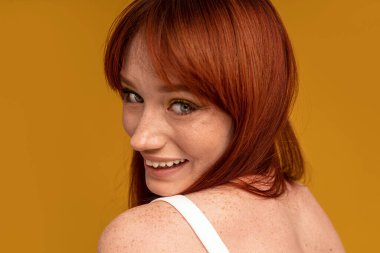 Mutlu kırmızı saçlı kadın kameraya bakıyor, sarı stüdyo arka planında poz veriyor. Gerçek insanların duyguları. Cilt bakımı.