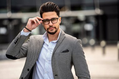Handsome young well-dressed man wearing fashionable eyeglasses, looking at the camera, posing outdoor. Businessman. Male model.