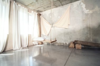 Modern beton tasarımcının fotoğrafı. Yastıklı ahşap salıncaklı. Doğal güneş ışığı. Duvarda süsleme var. Pencerelerde beyaz perdeler var. İlham.