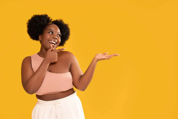 Happy woman with afro hair wearing sporty fashionable clothes and smiling, pointing on an empty background with finger