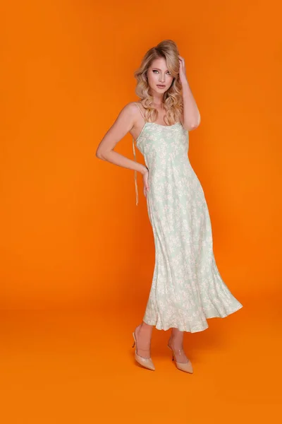 Blonde young woman in fashionable satin dress and high heels. Female model posing on an orange studio background in full length.