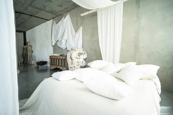 Natural style bedroom interior with a big bed, white linen and pillows. Concrete wall. Real photo of home design with fashionable decoration. Wooden furniture. Light curtains in cozy room