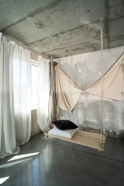Photo of modern concrete designer interior with wooden swing with pillows. Natural sunlight. Decoration on the wall. Long white curtains. Inspiration.