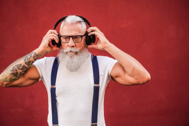 Handsome senior man with white beard, mustache and with tattoos listening to music with headphones. Concept of elderly people lifestyle and hobby. Real people. Red background. Copy space