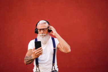 Handsome senior man with white beard, mustache and tattoos listening to music with headphones and mobile phone. Concept of elderly people lifestyle, hobby, technology. Real people. Red background. Copy space