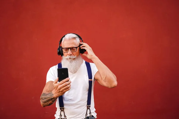 Handsome senior man with white beard, mustache and tattoos listening to music with headphones and mobile phone. Concept of elderly people lifestyle, hobby, technology. Real people. Red background. Copy space