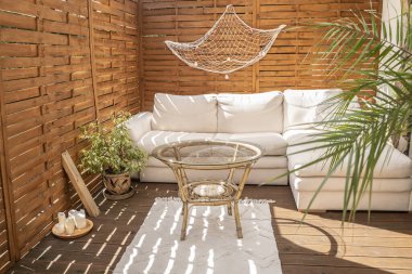 Real photo of relaxing boho zone in home. Wooden floor on terrace with comfy furniture and green plants. Decoration concept. Sunny summer day