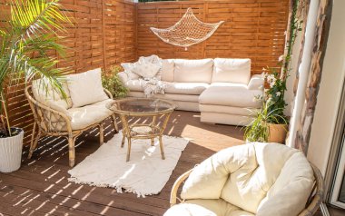 Real photo of relaxing boho zone in home. Wooden floor on terrace with comfy furniture and green plants. Decoration concept. Sunny summer day