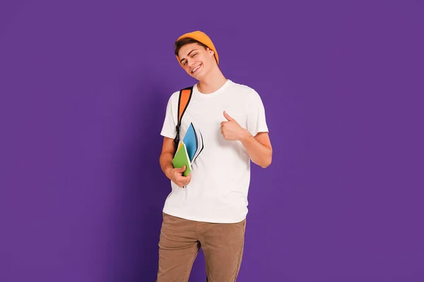 Young happy man student in casual clothes and cap, with backpack and notebooks, isolated on studio violet background. Boy showing okay sign. Education concept. Copy space