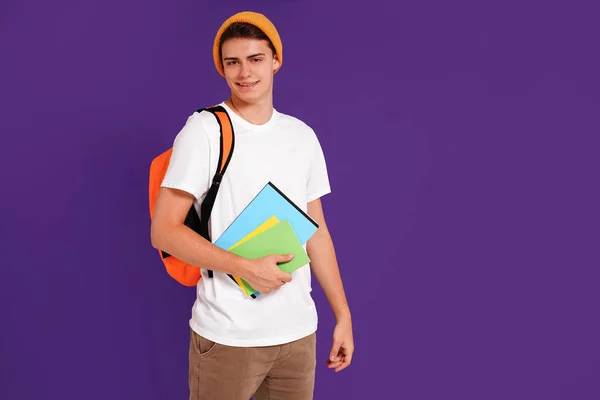 Young happy man student in casual clothes and cap, with backpack and notebooks, isolated on studio violet background. Education concept. Copy space
