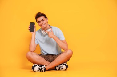 Happy smiling young man in casual clothes showing empty mobile phone screen, isolated on yellow studio background. People lifestyle concept. Copy space.