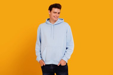 Young smiling handsome man in casual comfortable clothes posing isolated on yellow studio background, holding hands in pockets.. Real people emotions and lifestyle concept.