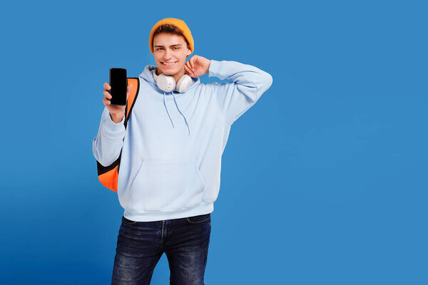 Photo of young fashionable student guy with cap and backpack smiling at camera, showing empty screen of mobile phone, isolated over blue studio background. A lot of copy space.