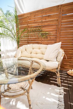 Real photo of relaxing boho zone in home. Wooden floor on terrace with comfy furniture and palm. Decoration concept. Sunny summer day.