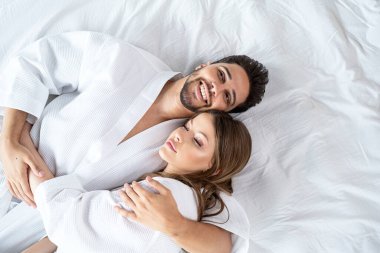 Young happy couple relaxing, lying in bed in white bathrobes, hugging and smiling. Morning light. Real people lifestyle.