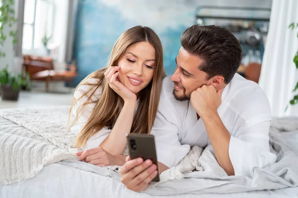 Happy Young Couple Relaxing Bed Home Bathrobes Using Mobile Phone — Stok fotoğraf