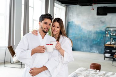 Young happy couple drinking coffee in the morning in white bathrobes,  enjoying a slow day together.  Relationship, love and family concept
