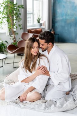 Beautiful young couple celebrating Saint Valentine's Day, spending romantic time together at home, wearing white bathrobes and hugging in bed. Real people lifestyle. Love.