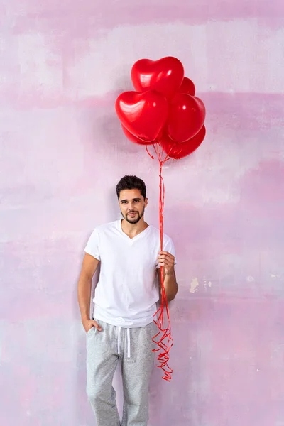 Handsome cute young man posing on the pink wall with red heart shaped balloons in hand, smiling and looking at camera. A lot of copy space. Valentines day. Love.