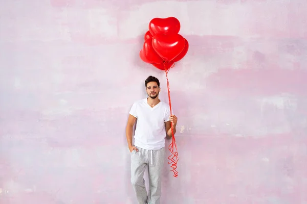 Handsome cute young man posing on the pink wall with red heart shaped balloons in hand, smiling and looking at camera. A lot of copy space. Valentines day. Love.