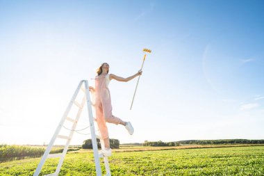 Merdiven üzerinde fırçayla gülümseyen bir kadının kavramsal fotoğrafı. Gökyüzünü maviye boyayan mutlu kız. Yaz titreşimleri. Doğa aşığı. Reklamınız için çok fazla kopya alanı var.