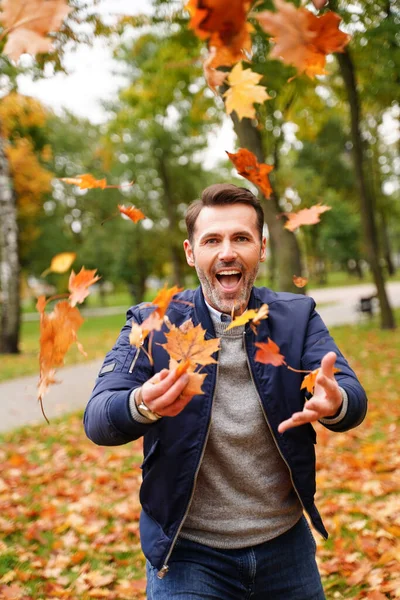행복하게 남자가 황금빛 공원에서 나뭇잎을 가지고 놀면서 웃으며 카메라를 봅니다 — 스톡 사진