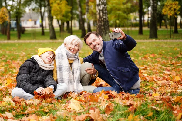 幸せなお母さん お父さんと息子は一緒に楽しい時間を過ごし 秋のゴールデンパークで携帯電話で自撮りをします 本当の家族のライフスタイルの概念 — ストック写真