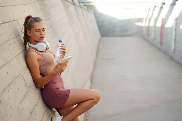 Young fit blonde woman after training, using mobile phone, scrolling social media and relaxing. Outdoor photo. Healthy lifestyle.