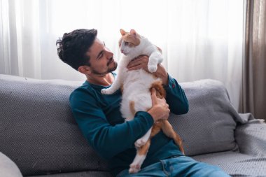 Gri bir kanepede oturan genç adam kollarında kahverengi beyaz bir kedi tutuyor.