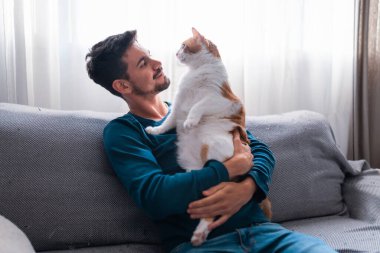 Gri bir kanepede oturan genç adam kollarında kahverengi beyaz bir kedi tutuyor.