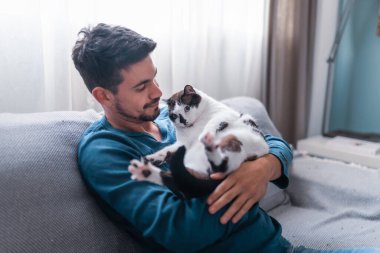 Gri bir kanepede oturan genç adam kollarında mavi gözlü siyah beyaz bir kedi tutuyor.