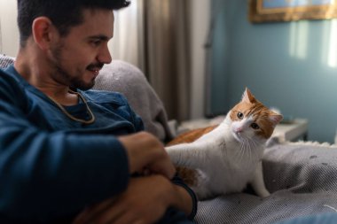 Genç adam kanepede kahverengi ve beyaz bir kediyle oynuyor. Kapat.