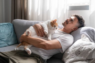 Kanepede oturan genç adam kahverengi ve beyaz kediye sarılıyor.