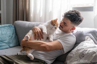 Genç adam kanepede uzanıyor, kahverengi ve beyaz bir kediye sarılıyor.