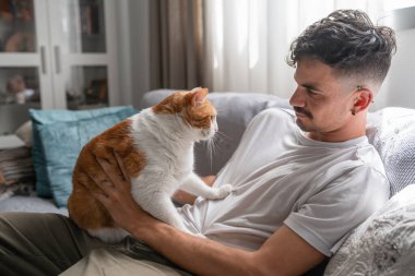 Genç adam kanepede uzanıyor, kahverengi ve beyaz bir kediye sarılıyor.