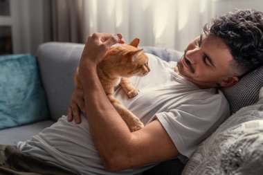 Kanepede uzanmış genç bir adam kahverengi bir tekir kediyle etkileşime geçiyor.