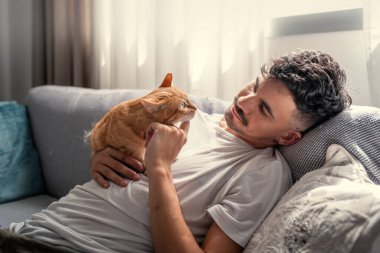 Kanepede uzanmış genç bir adam kahverengi bir tekir kediyle etkileşime geçiyor.