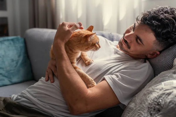 Kanepede uzanmış genç bir adam kahverengi bir tekir kediyle etkileşime geçiyor.