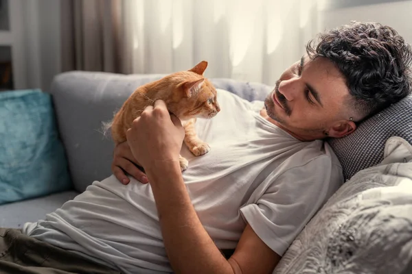 Kanepede uzanmış genç bir adam kahverengi bir tekir kediyle etkileşime geçiyor.